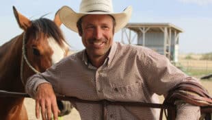 A smiling man in a cowboy hat leaning on a fence with a horse behind him.