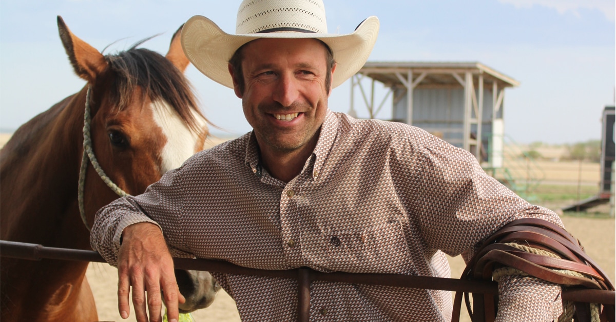 A smiling man in a cowboy hat leaning on a fence with a horse behind him.